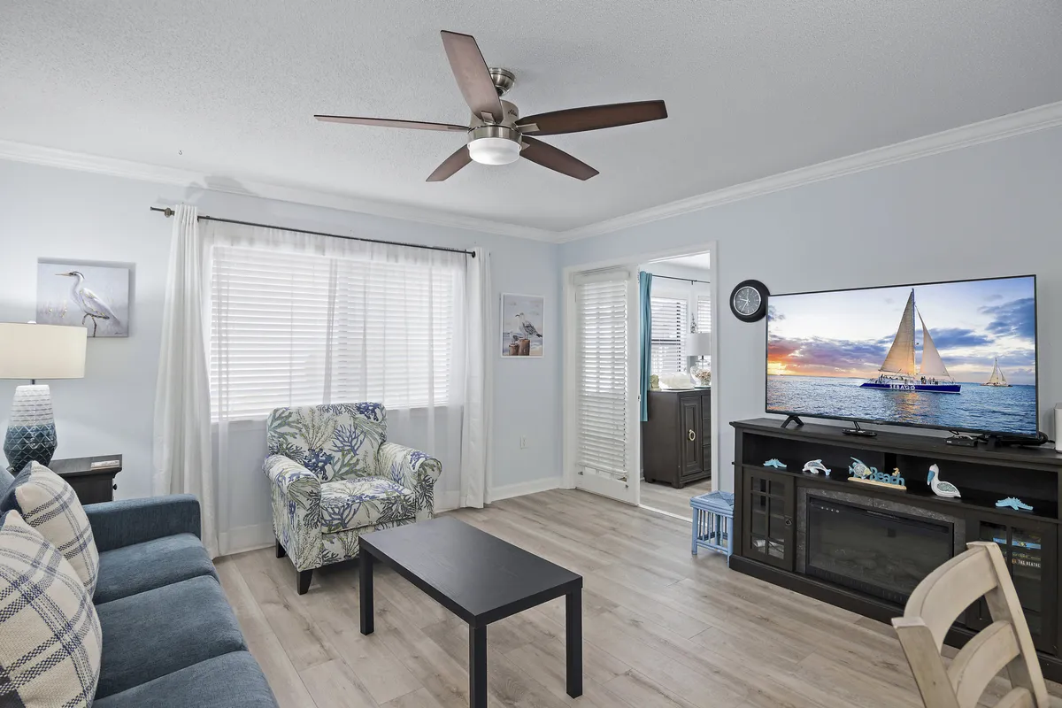 The image showcases a cozy, coastal-inspired living room featuring a plush blue sofa with plaid cushions, a comfortable armchair with a beachy pattern, and a wide-screen TV displaying a tranquil sailboat scene. The decor includes a dark wood media console adorned with beach-themed accents, all set in a sunlit, inviting space perfect for relaxing after a day at the beach.