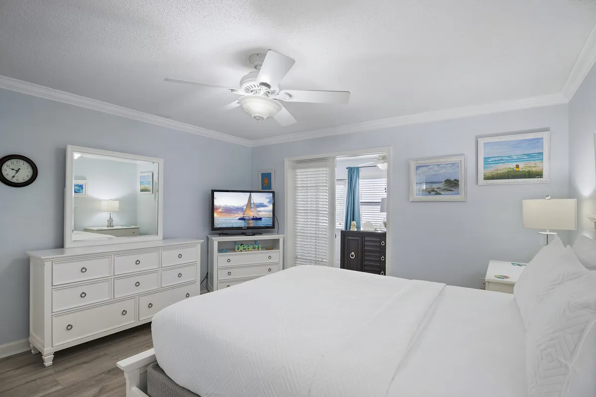 A bright, serene primary bedroom featuring a large white bed with crisp bedding, a white dresser with mirror, and a television displaying a tranquil sailboat scene. Subtle beach-inspired wall art and soft lighting create a restful, inviting ambiance.