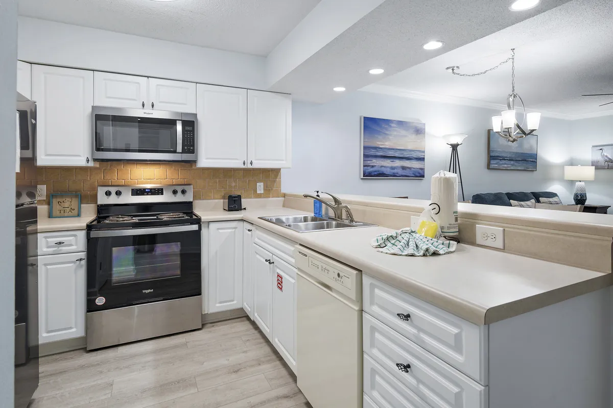 An open kitchen view with stainless steel stove and microwave, white cabinetry and countertops, and a double sink with faucet. The kitchen opens into a living area adorned with wall art, underscoring the open-plan layout and the integration of cooking and social spaces.