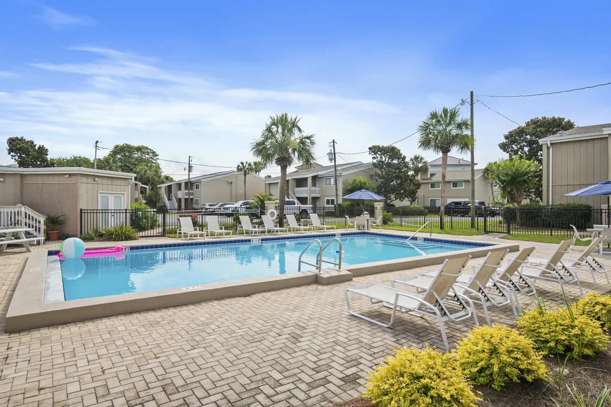A spacious, seasonally heated community swimming pool surrounded by palm trees, poolside lounge chairs, and adjacent condo buildings. The area is sun-drenched and inviting, offering plenty of space for swimming, sunbathing, and casual socializing, with easy access from the condo to recreational amenities.