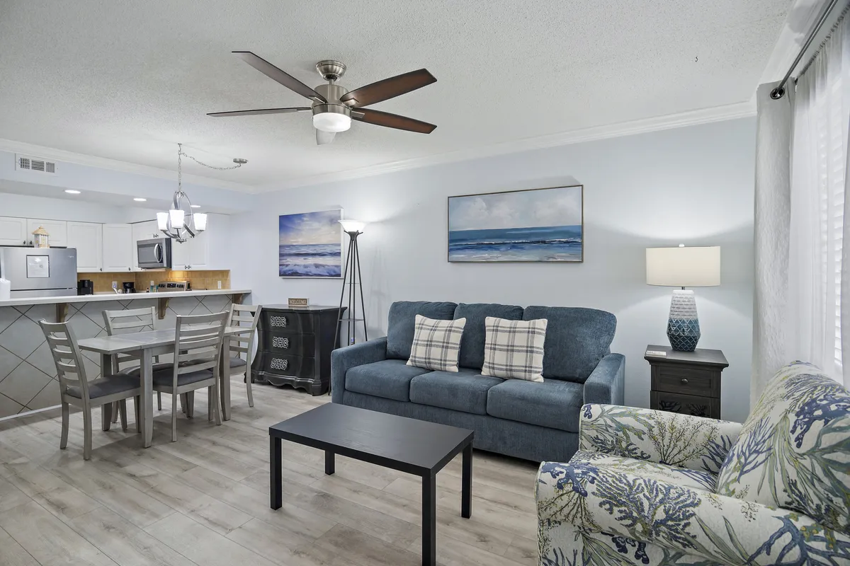 A welcoming open-plan living room featuring a blue sofa with plaid pillows, a coastal-themed armchair with coral pattern, and a dining area adjacent to a bright kitchen with white cabinetry and appliances. The space is filled with natural light, evoking a calm and relaxed atmosphere perfect for unwinding after beach outings or gathering with family and friends.