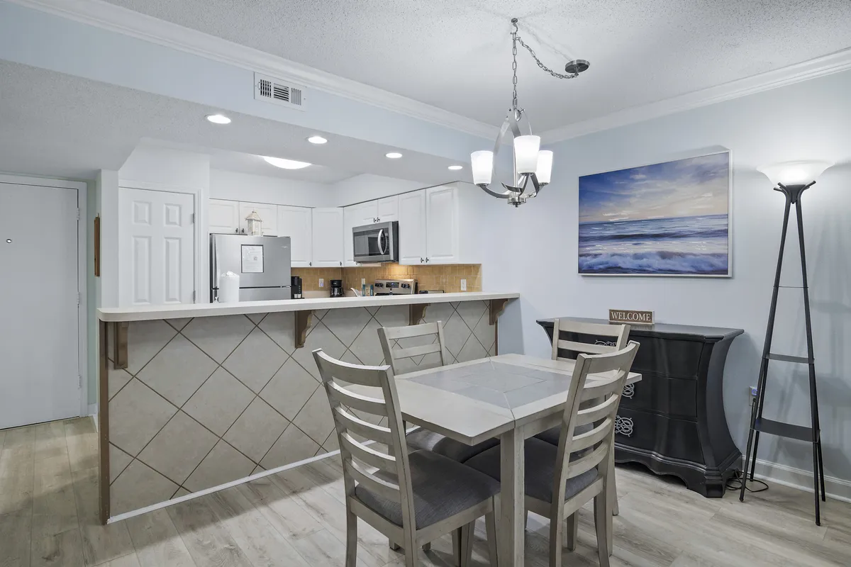 This image features a dining table with four chairs situated next to a kitchen with white cabinetry and stainless steel appliances. Decorative ocean-themed artwork and a unique black chest with a welcome sign add a touch of personality. The open layout provides a warm, communal feel, ideal for group meals and gatherings.