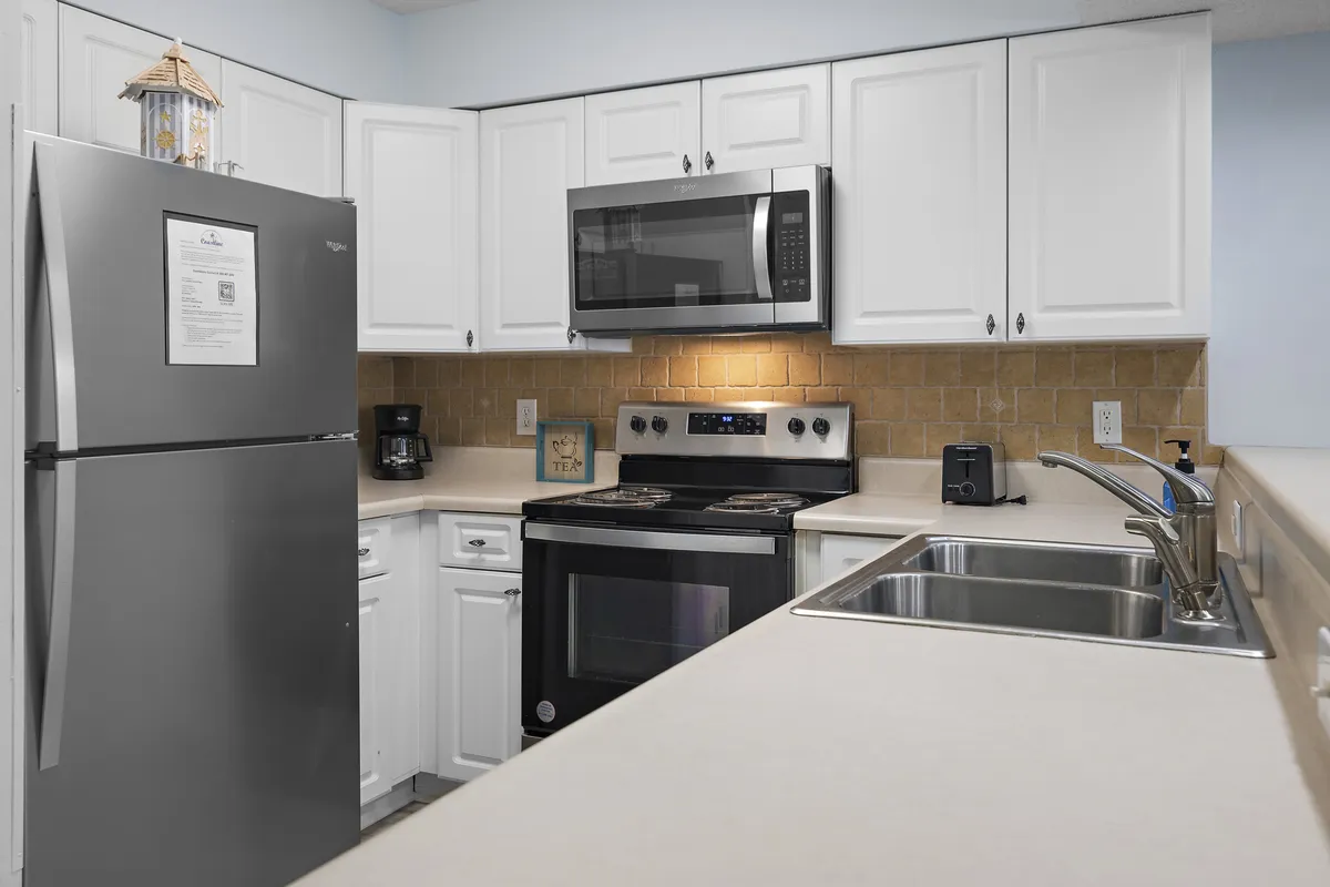 A practical kitchen scene showcasing a stainless steel refrigerator, electric stove with oven, microwave oven, and double sink. The layout is efficient, with plenty of workspace for meal prep or morning routines like coffee making.