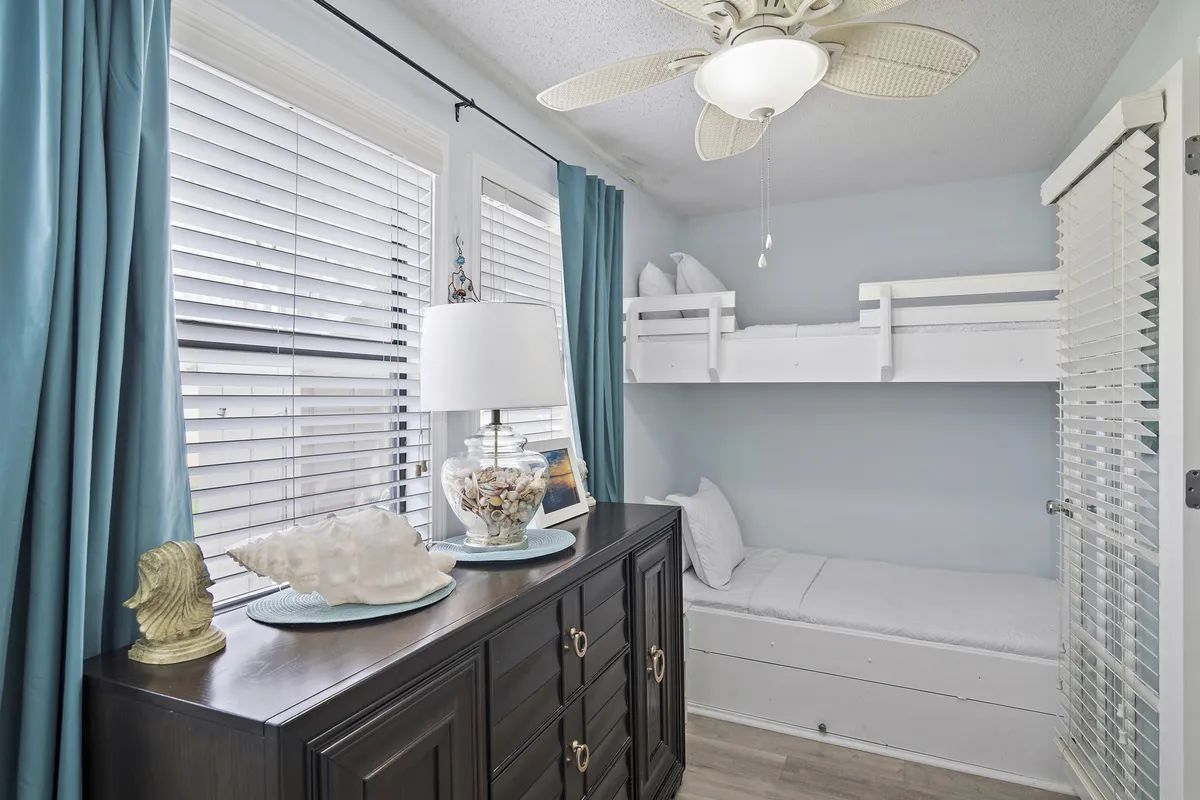 A cozy bunk nook with white twin-over-twin bunk beds, a dark wooden dresser topped with a decorative seashell lamp, and large windows draped in teal curtains. Natural light fills the small, cheerful space, making it ideal for children.