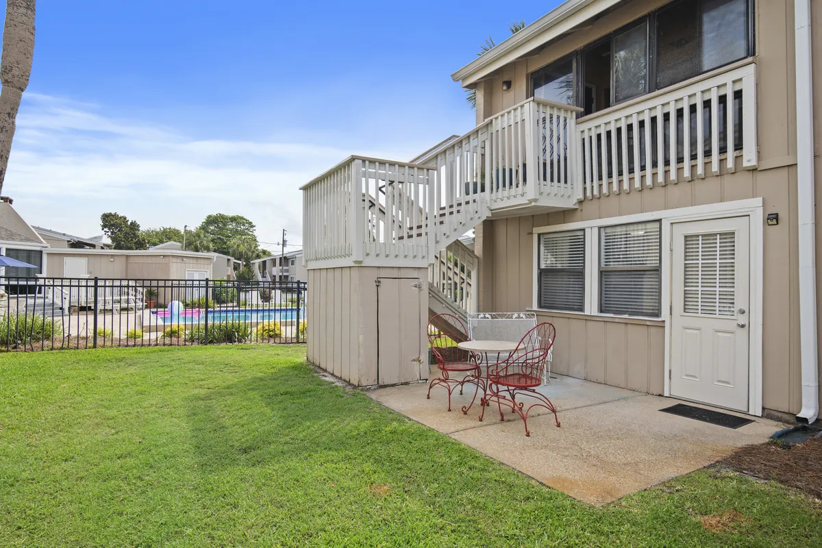 A cozy ground-floor patio with a red metal table and chairs overlooks a well-maintained green lawn and the community pool beyond a fence. The space feels welcoming for families and pets, encouraging outdoor dining or relaxation after a day of activities.