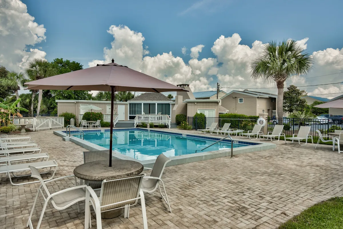 The photo captures a calm, inviting community pool surrounded by palm trees, lounge chairs, and a round patio table with an umbrella. The landscaped setting suggests a relaxing communal space where guests can unwind after walks or playtime with their pets.