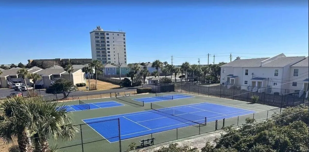 Vibrant blue tennis and pickleball courts surrounded by palm trees and residential buildings. A nearby bench provides a spot for spectators or players to rest, emphasizing the active recreation opportunities on site.