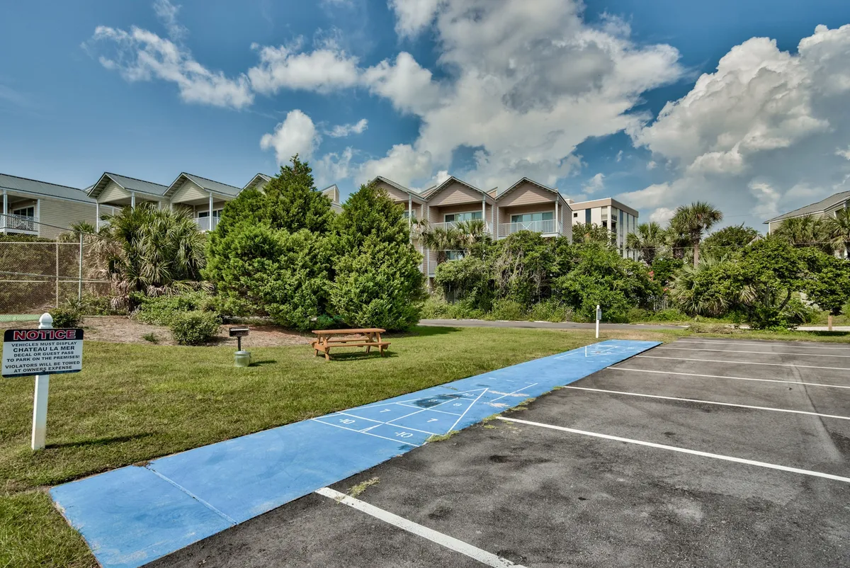 This exterior image displays a blue shuffleboard court and a picnic table set on a grassy area, surrounded by condo buildings and adjacent parking. The scene is relaxed and open, highlighting green spaces that are ideal for pet-friendly walks and outdoor play. The well-maintained common areas are visible, offering a sense of community and leisure for guests and their pets.