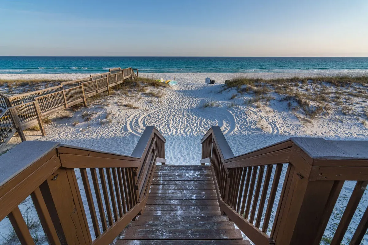 A peaceful scene featuring a wooden staircase descending to a pristine white sandy beach, with blue ocean horizons and a row of beach chairs and umbrellas already set up for guests. The setup reflects the daily preparations awaiting residents and visitors.
