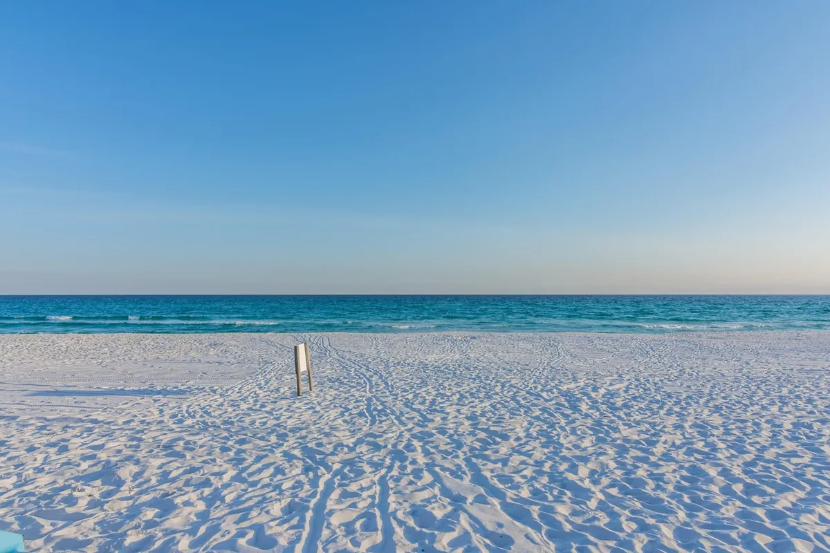 This image captures a pristine stretch of white sand beach under a clear blue sky, with a calm turquoise ocean and a beach access sign. The tranquil scene invites viewers to imagine walking hand-in-hand along the shoreline or relaxing with daily chair and umbrella setups.