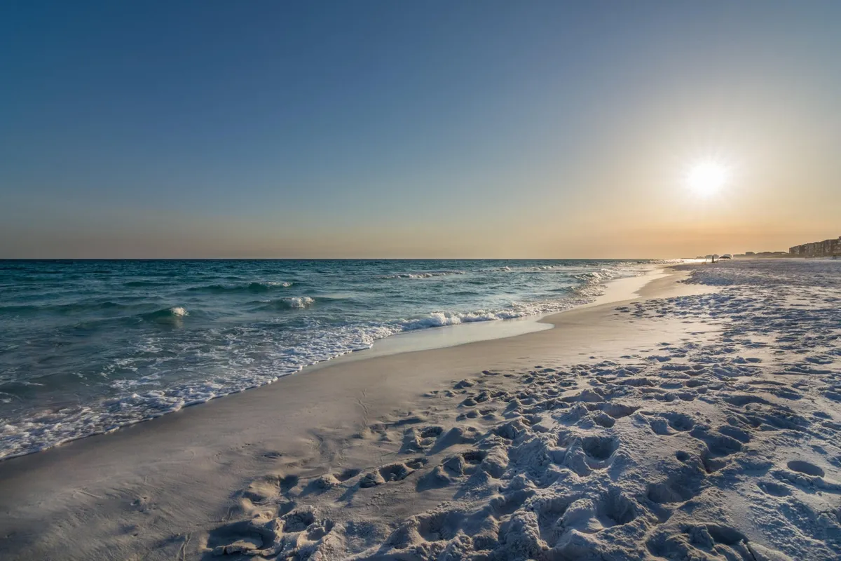 A serene expanse of sandy beach with gentle ocean waves, the sun low in the sky casting a warm glow, and distant shoreline buildings. Footprints in the sand and a sense of inviting calmness capture the quintessential beach experience described in the section.