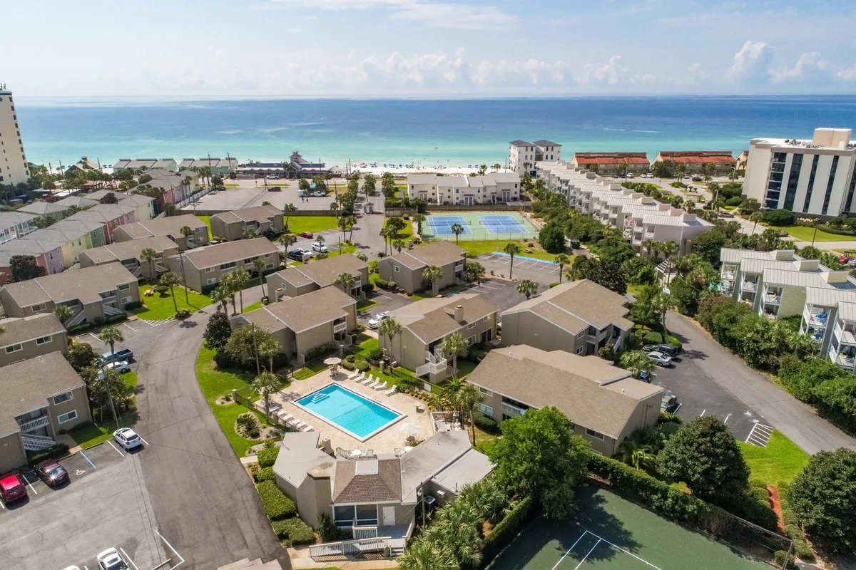A vibrant exterior shot capturing the central heated community pool surrounded by lounge chairs, adjacent tennis and pickleball courts, and the private beach access visible in the background. Ground-floor condo units with patios cluster around the amenities, illustrating the close proximity of all resort-style perks.