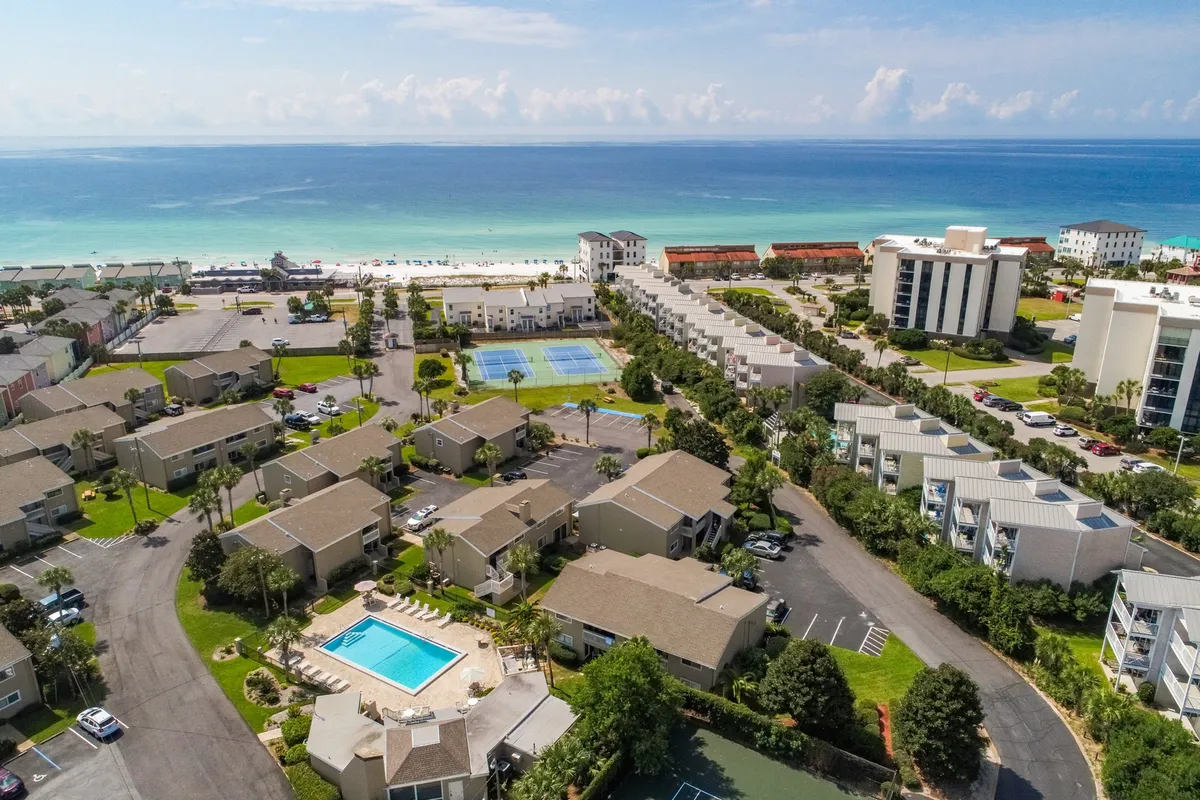 A wide aerial view highlights the heated community pool with lounge chairs, tennis and pickleball courts, private beach access with umbrellas and chairs, and a cluster of condo buildings. The image showcases multiple outdoor amenities and the proximity of everything guests might want.