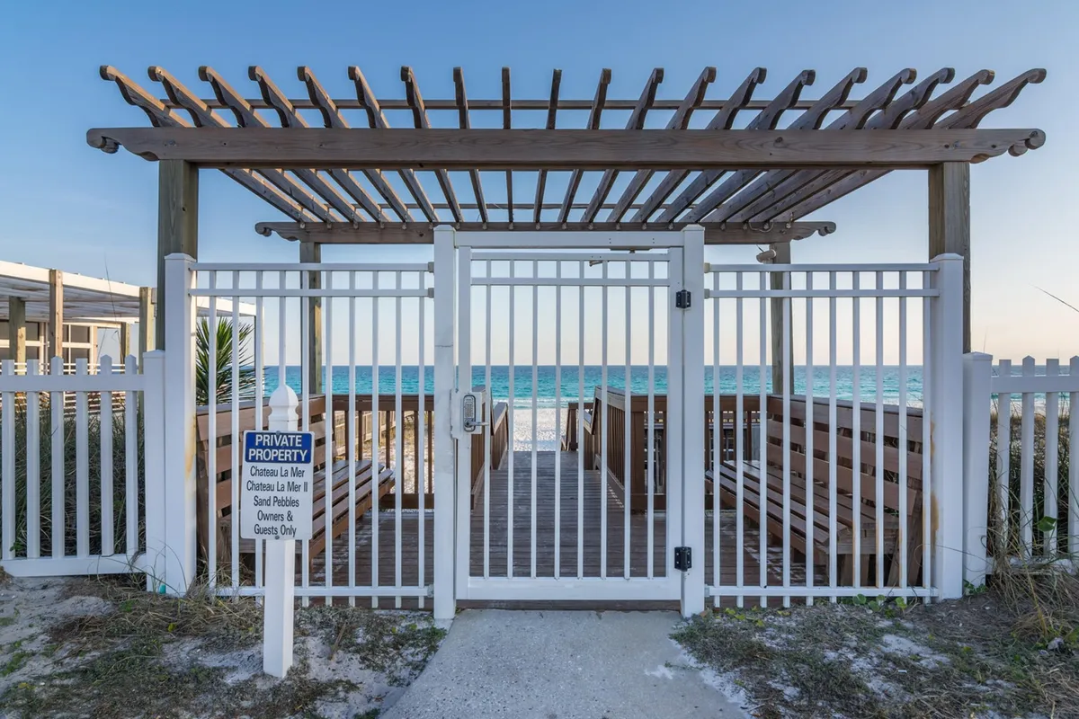 A white wooden gate opens to a pergola structure that perfectly frames a stunning ocean view. The foreground features a sandy pathway inviting guests toward the beach, while the open sky and tranquil water set a romantic, serene backdrop. This spot is ideal for enjoying sunsets, intimate gatherings, or simply soaking up the coastal ambiance.