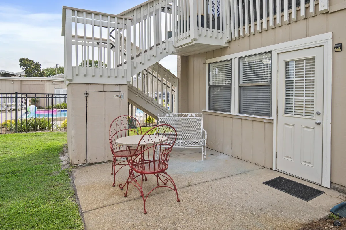 The photo shows an exterior patio with red metal chairs and a small round table, set under a spiral staircase. The area is ideal for enjoying fresh breezes, sipping morning coffee, or reading a book while overlooking the outdoors.