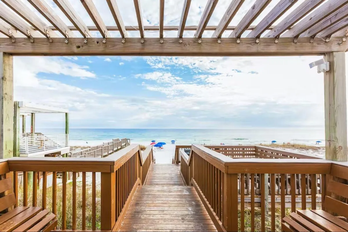 A picturesque exterior shot of a wooden walkway leading directly to a pristine beach, dotted with colorful umbrellas set up and ready for guests. The calm blue sky and expansive beach invite visitors to imagine their private setup and leisure time.